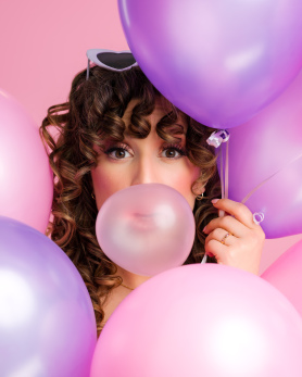 Bethany blowing a bubblegum bubble with pink and purple balloons